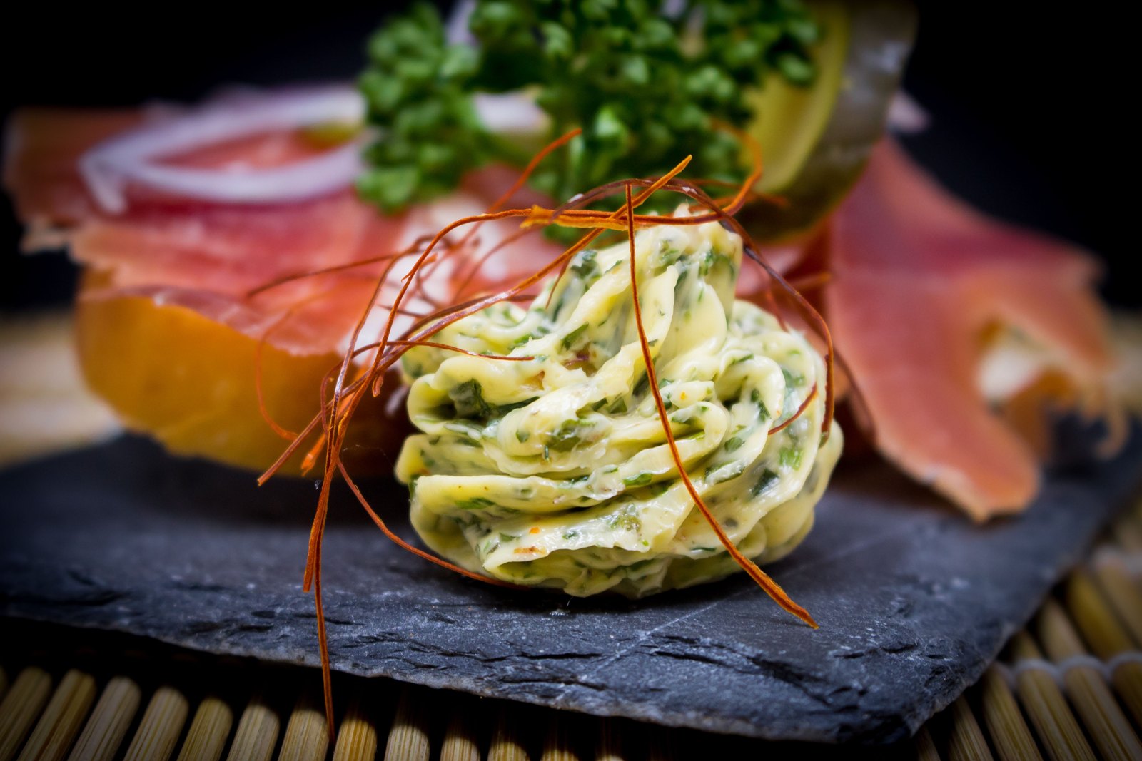 A piped curl of pale herb butter on a dark slate board beside toasted bread and thin slices of cured meat