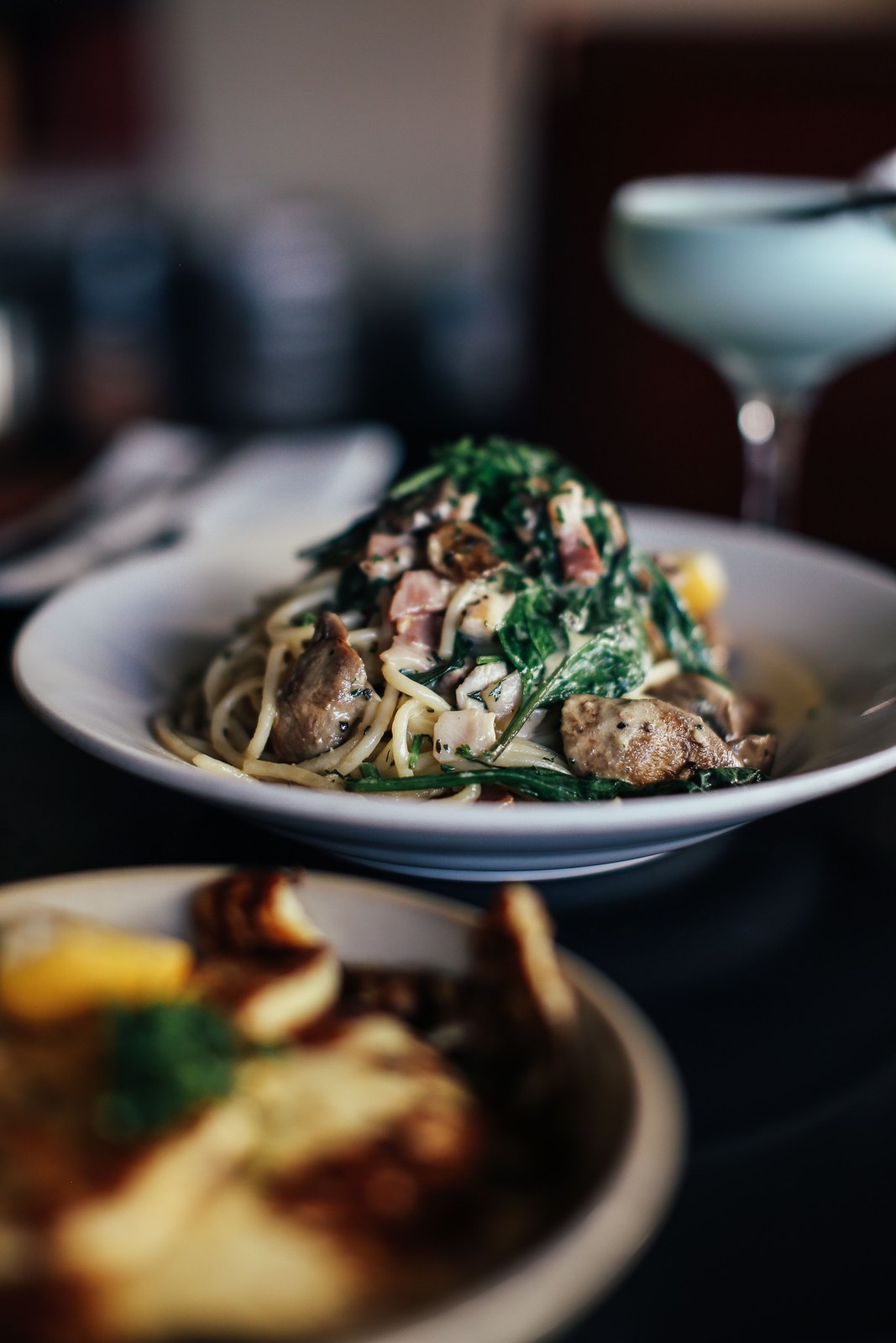 A deep white bowl of spaghetti tossed with greens and herbs, photographed on a dark restaurant table
